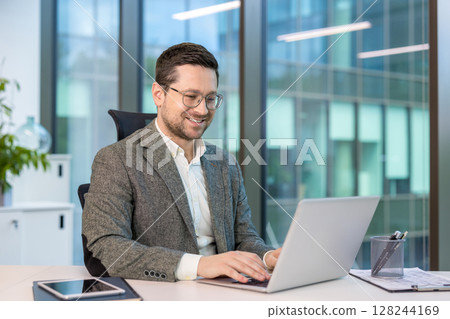 Smiling young businessman sitting in a suit in the office at a table and working on a laptop. Smiling young businessman sitting in a suit in the office at a table and working on a laptop. 128244169