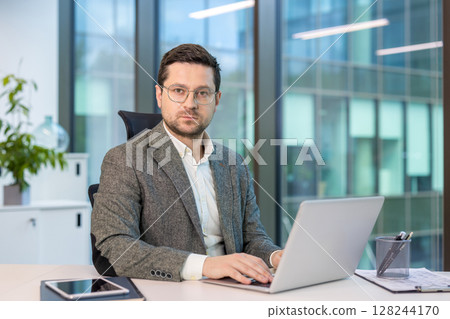 Portrait of a serious and confident young man in a suit and glasses sitting in the office at a desk, working on a laptop and looking at the camera. Portrait of a serious and confident young man in a suit and glasses sitting in the office at a desk, working on a laptop and looking at the camera. 128244170
