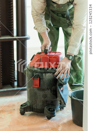 Construction Worker with Red Shop Vacuum Cleaner, Interior Remodeling, Dust Control, Wood Floors, Green Overalls, Workwear, Industrial Setting Construction Worker with Red Shop Vacuum Cleaner, Interior Remodeling, Dust Control, Wood Floors, Green Overalls, Workwear, Industrial Setting 128245334