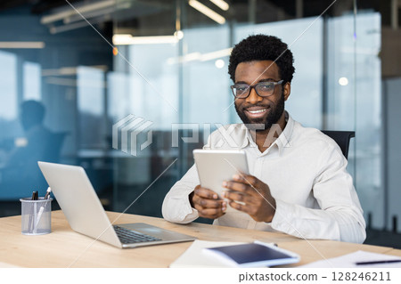 A smiling African American man in glasses uses a tablet at his office desk, a laptop sits nearby. 128246211