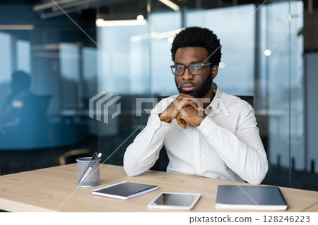 A thoughtful African-American man wearing glasses and a white shirt sits at his desk in a modern office setting. A thoughtful African-American man wearing glasses and a white shirt sits at his desk in a modern office setting. 128246223