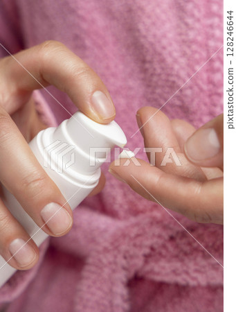 Woman in pink bathrobe pumping cream on finger and holding a cosmetic bottle closeup Woman in pink bathrobe pumping cream on finger and holding a cosmetic bottle closeup 128246644