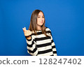 A beautiful Hispanic teenager in her 15s wearing a striped sweater posing isolated against a blue background. Lifestyle concept. Rubbing her fingers together, gesturing for money 128247402