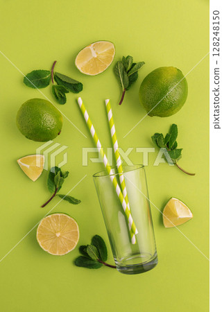 Preparing lemonade. Limes, mint leaves, straws, and a glass on a vibrant green background. Preparing lemonade. Limes, mint leaves, straws, and a glass on a vibrant green background. 128248150