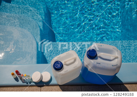 Top view of pool maintenance supplies including chlorine tablets, test kit bottles, and large chemical canisters arranged on the edge of a clean blue pool. 128249188