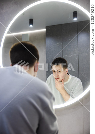 Young man inspects his face in the mirror with a concerned look, gently touching his chin in a modern bathroom during his morning routine. Young man inspects his face in the mirror with a concerned look, gently touching his chin in a modern bathroom during his morning routine. 128249216
