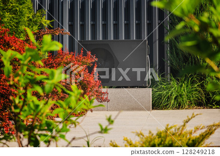 Outdoor heat pump unit installed near a modern building, surrounded by decorative plants and colorful shrubs on a sunny day 128249218