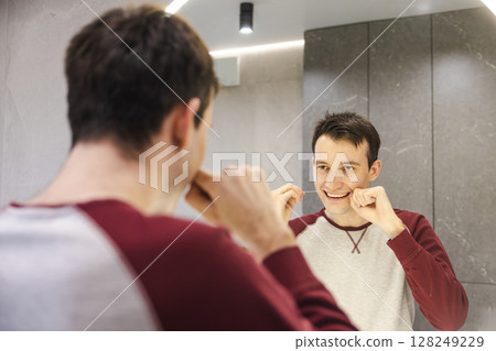 Young man flosses his teeth in front of a mirror, smiling with focus as he follows a healthy oral care routine to maintain dental hygiene and cleanliness. 128249229