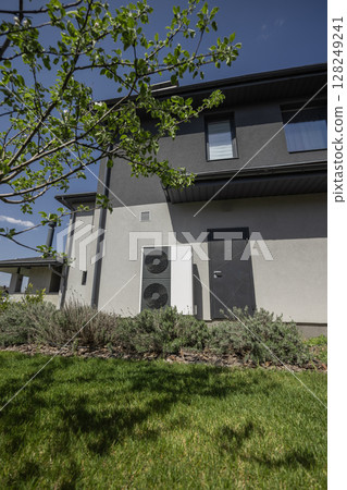 Modern heat pump system mounted on residential house wall surrounded by green garden and spring trees 128249241
