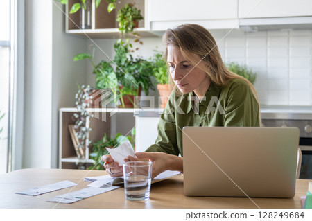 Pensive sorrowful woman reviewing financial documents with laptop and water glass at home kitchen 128249684