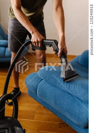 Close-up vertical cropped shot of cleaning service worker using washing vacuum cleaner to clean blue sofa in modern living room. Concept of hygiene and cleanliness in domestic environment. 128249918