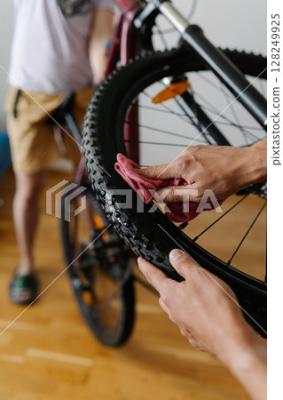 Vertical cropped shot of cyclist cleaning tire of bicycle wheel with cloth, ensuring optimal performance and maintaining bike pristine condition in workshop, close-up. Concept of bike maintenance. Vertical cropped shot of cyclist cleaning tire of bicycle wheel with cloth, ensuring optimal performance and maintaining bike pristine condition in workshop, close-up. Concept of bike maintenance. 128249925
