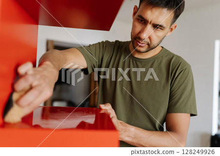Portrait of young focused man cleaning wiping red shelf surface with sponge and detergent ensuring spotless and hygiene at home, close-up. Concept of cleanliness in domestic environment. 128249926