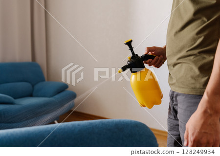 Man spraying water onto blue sofa using yellow plastic sprayer, diligently cleaning furniture in cozy living room at home, ensuring hygiene and freshness, close-up. Concept of hygiene and cleanliness. 128249934