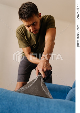 Close-up vertical portrait of man in causal clothing using washing vacuum cleaner to clean blue sofa in home setting, demonstrating modern approach to household chores and upholstery maintenance. Close-up vertical portrait of man in causal clothing using washing vacuum cleaner to clean blue sofa in home setting, demonstrating modern approach to household chores and upholstery maintenance. 128250168