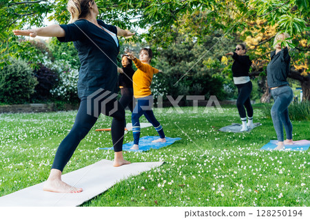 Female yoga class in park. Group of diverse women doing stretching pose exercising together with instructor on green grass lawn. Sport activity for health, wellbeing, mental health. Selective focus 128250194