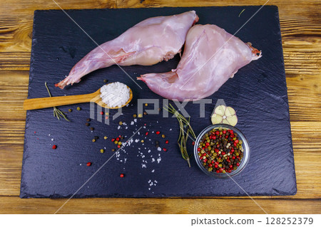Raw rabbit legs with spices on a black slate cutting board on wooden table. Top view Raw rabbit legs with spices on a black slate cutting board on wooden table. Top view 128252379
