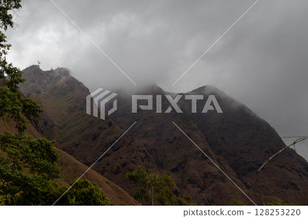 Dark cloudy sky hangs over rugged green and brown mountain landscape. Dark cloudy sky hangs over rugged green and brown mountain landscape. 128253220