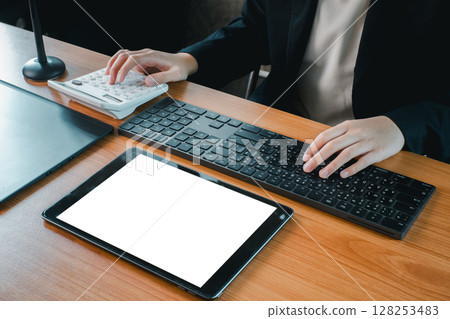Business professional using a tablet, keyboard, and calculator at a wooden desk in a modern office setting. 128253483