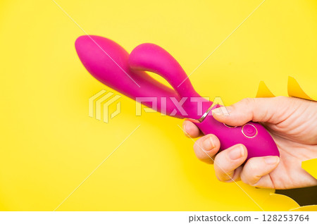 Female hand with magenta vibrator sticking out from yellow paper background. Copy space. Female hand with magenta vibrator sticking out from yellow paper background. Copy space. 128253764