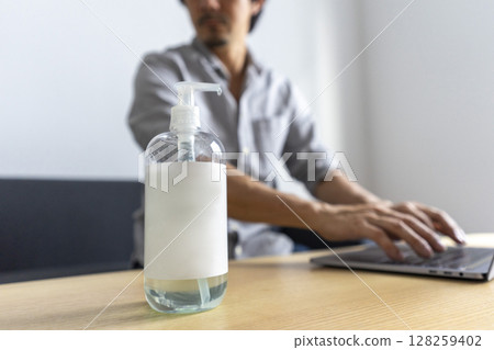 Hand sanitizer gel standing on wooden table. Adult Asian man cleaning hand with sanitizer gel while using laptop computer. 128259402