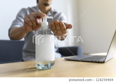 Hand sanitizer gel standing on wooden table. Adult Asian man cleaning hand with sanitizer gel while using laptop computer. 128259404