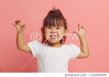 Beautiful little Asian girl has fun and shows her tongue by raising her hands up while standing on a pink isolated. Beautiful little Asian girl has fun and shows her tongue by raising her hands up while standing on a pink isolated. 128260699