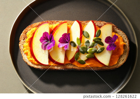 A beautifully arranged toast features creamy pumpkin spread topped with vibrant apple slices and scattered pumpkin seeds, presented on a clean white plate 128261622