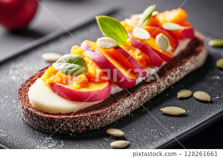A beautifully arranged toast showcases vibrant slices of apple and peach, topped with creamy spread and seeds, resting on dark bread. The presentation highlights fresh ingredients 128261661