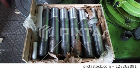 A crate of newly manufactured flange studs, steel threaded rods, with industrial machinery in the background. Close-up of metal flange studs, packaged in a wooden box for transport. Metal studs  128262891