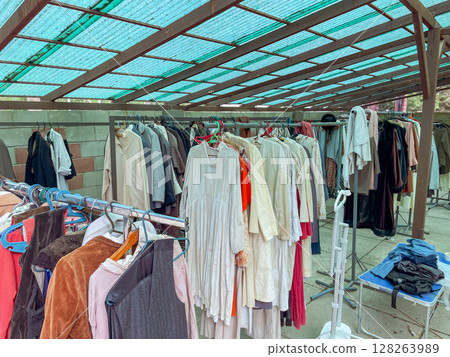 A diverse array of vintage clothing hangs on metal racks under a teal canopy. Garments include dresses, coats, and shirts in various colors and textures. A diverse array of vintage clothing hangs on metal racks under a teal canopy. Garments include dresses, coats, and shirts in various colors and textures. 128263989