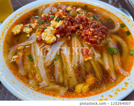 A close-up of a steaming bowl of noodles in a rich, red broth, topped with minced garlic, green onions, and a dollop of spicy chili paste, showcasing a hearty meal. 128264007