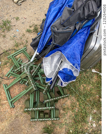 A chaotic arrangement of blue and black umbrellas lies atop green metal stands on a gravel surface, with scattered grass and droplets of water visible. A chaotic arrangement of blue and black umbrellas lies atop green metal stands on a gravel surface, with scattered grass and droplets of water visible. 128264008