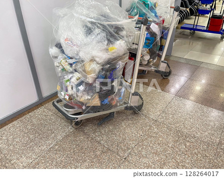 A cart filled with a large, transparent plastic bag overflowing with assorted waste items, including bottles and packaging, positioned on a tiled floor. 128264017