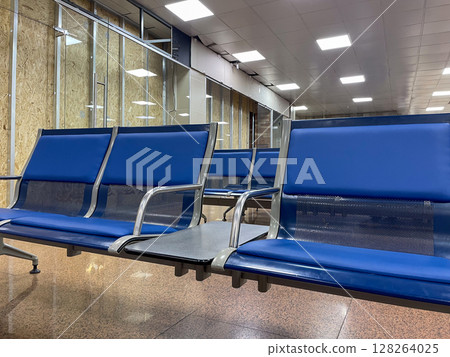 The image showcases a row of sleek blue airport seats with metallic frames, positioned in a spacious terminal. The polished floor reflects overhead lights. The image showcases a row of sleek blue airport seats with metallic frames, positioned in a spacious terminal. The polished floor reflects overhead lights. 128264025