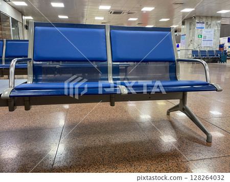 Two blue metal-framed airport seats are positioned in a spacious terminal. The floor features a glossy, speckled pattern, while the background shows empty waiting areas. Two blue metal-framed airport seats are positioned in a spacious terminal. The floor features a glossy, speckled pattern, while the background shows empty waiting areas. 128264032