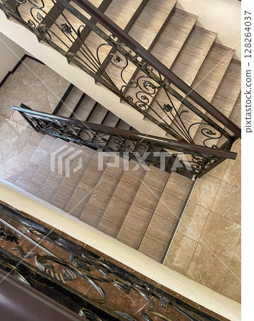 A top-down view of a staircase featuring intricate wrought iron railings. The steps are light-colored tiles, contrasting with the dark railing and wooden accents. 128264037
