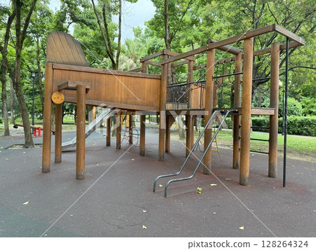 Large wooden play structure featuring a slide and climbing frame resting on a soft rubberized surface in a park setting, surrounded by trees and greenery for children's playtime Large wooden play structure featuring a slide and climbing frame resting on a soft rubberized surface in a park setting, surrounded by trees and greenery for children's playtime 128264324