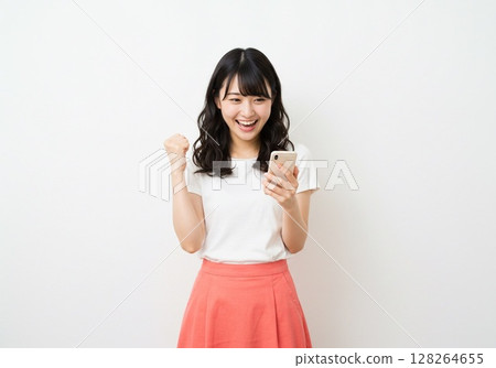 A Japanese woman posing in a fist pump while looking at her smartphone screen (white background, for cutting) 128264655