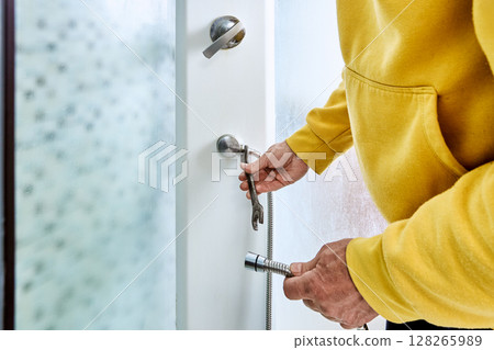 Tightening connection nut with open end wrench, plumber attaches new shower hose to water outlet fitting on panel inside shower cabin. 128265989