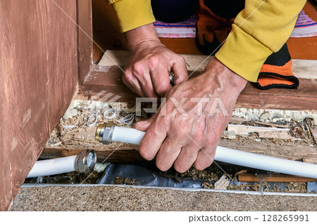 Plumber uses hands to connect multilayer pipe with compression fitting, working on plumbing system near wooden floor and wall. 128265991