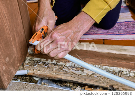 Plumber uses hand pipe cutter to cut PEX pipe near wooden floor, preparing pipe end for connection during plumbing work in residential building. 128265998