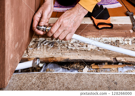 Person connects compression fitting to white metal composite pipe near damaged wooden floor. 128266000