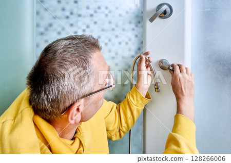 Inside shower cabin connector nut of flexible shower hose is being hand tightened by plumber onto water outlet on shower panel surface. 128266006