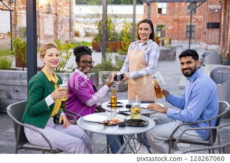 A female waiter processes payment with a portable terminal for a customer at an outdoor restaurant. 128267060