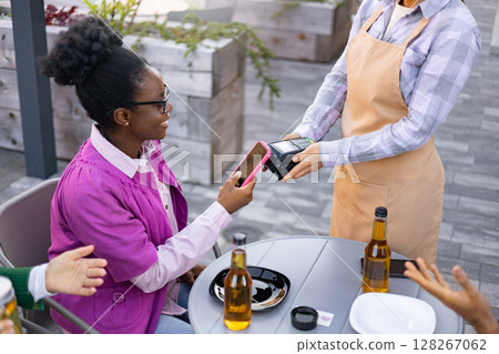 A female waiter using a portable terminal, assisting a customer with a payment in the outdoor cafe. 128267062