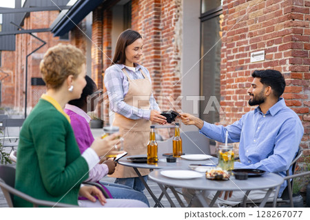 A female waiter in an apron hands a portable terminal to a customer at a restaurant table for payment. 128267077