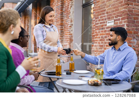 A female waiter is at a table and is asking a customer to pay the bill using a portable payment terminal. Other customers are present. 128267088