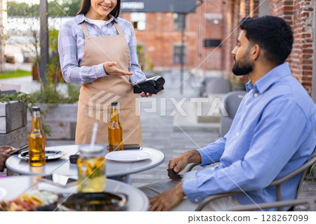 A female waiter offers a payment terminal to a customer at an outdoor restaurant table. 128267099