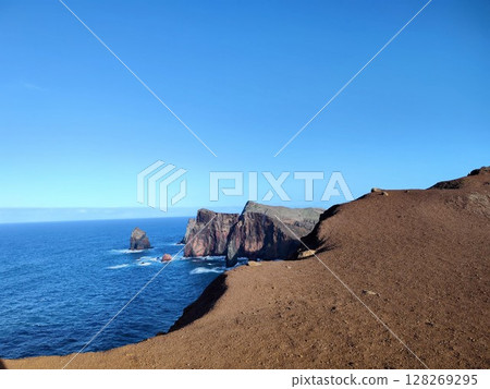 Rugged cliffs rise along a vibrant blue ocean, with waves gently breaking against the rocky formations. Madera, Portugal Rugged cliffs rise along a vibrant blue ocean, with waves gently breaking against the rocky formations. Madera, Portugal 128269295